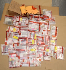 A large collection of sealed evidence bags spread out on a table, each containing various items with labels and red seals Brown paper bags are visible in the background