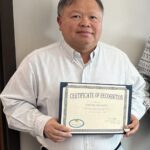 A person wearing a striped shirt holds a framed Certificate of Recognition