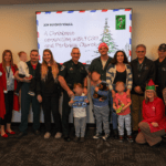 A group of adults and children pose together indoors at a Christmas themed event The background features a festive presentation slide titled "Joy Beyond Walls " Some people hold toys, and several are dressed in holiday attire