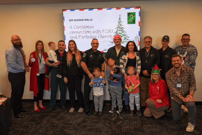 A group of adults and children pose together indoors at a Christmas themed event The background features a festive presentation slide titled "Joy Beyond Walls " Some people hold toys, and several are dressed in holiday attire