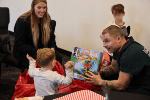 A man and a woman are sitting with two children The man is handing a boxed toy to the toddler, who is excitedly reaching for it There are red bags on the floor, and they all appear to be in a cheerful setting