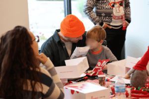 A group of people sitting around a table with fast food boxes A person wearing an orange beanie is holding a child closely The table is covered with festive decorations and food Another person is standing in a holiday sweater in the background