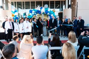 A group of people, including some in white lab coats, stand in front of a building's entrance decorated with blue and white balloons A ribbon cutting ceremony is taking place Onlookers are seated, capturing the event