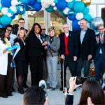 A group of people, including a woman cutting a ribbon, at a building's entrance They are surrounded by blue, white, and gold balloons Some are clapping, and others are wearing lab coats People are capturing the moment with phones