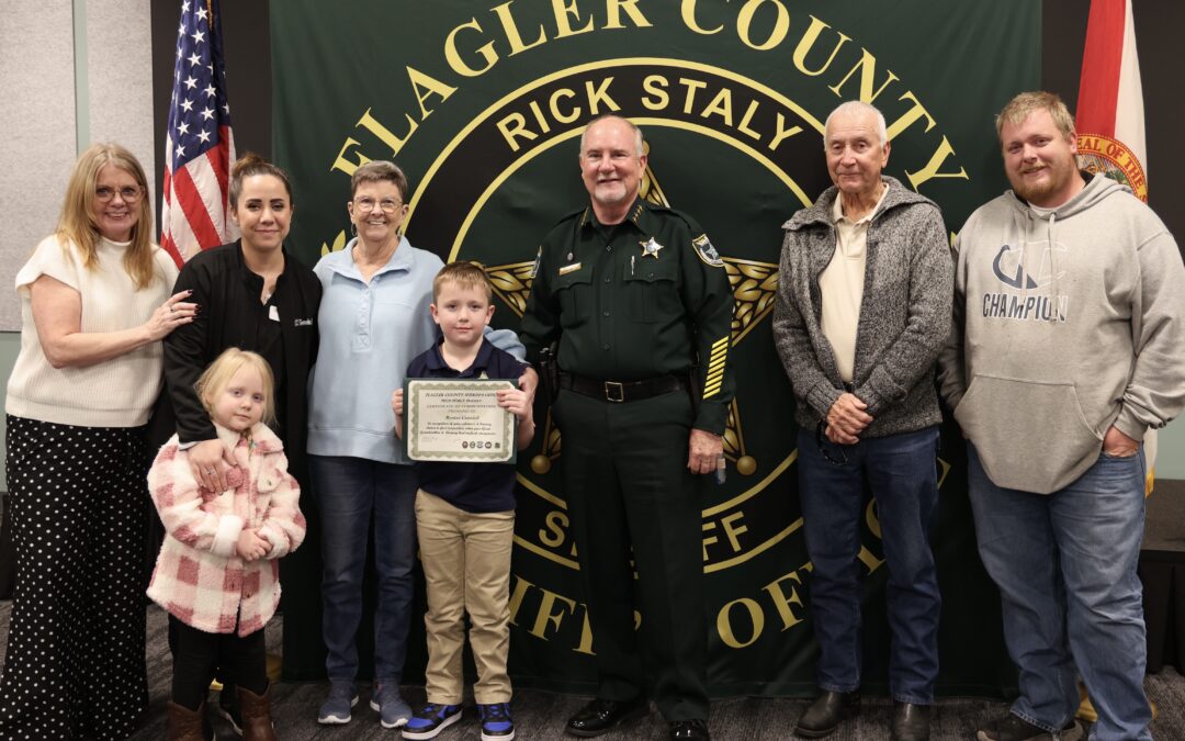 A group of people, including a sheriff, stand in front of a Flagler County Sheriff's Office banner A boy holds a certificate, and an American flag is in the background The group appears to be in a celebratory or award giving setting