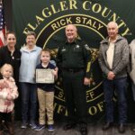 A group of people, including a sheriff, stand in front of a Flagler County Sheriff's Office banner A boy holds a certificate, and an American flag is in the background The group appears to be in a celebratory or award giving setting