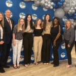 A group of nine people, both men and women, stand smiling in front of a backdrop with a logo and balloons They are dressed in business or semi casual attire, and the setting appears to be a formal event or ceremony