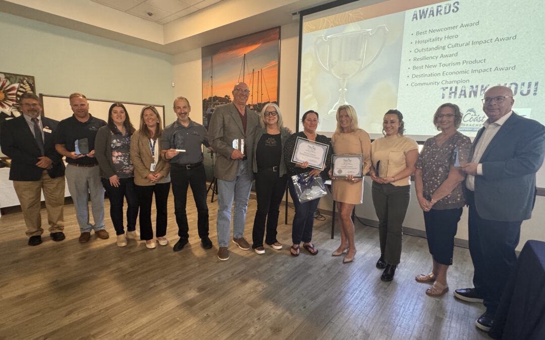 A group of people stand in a room holding awards and certificates A presentation screen in the background displays "Awards" and "Thank You" along with various award titles The floor is wooden, and there are scenic pictures on the wall