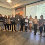 A group of people stand in a room holding awards and certificates A presentation screen in the background displays "Awards" and "Thank You" along with various award titles The floor is wooden, and there are scenic pictures on the wall