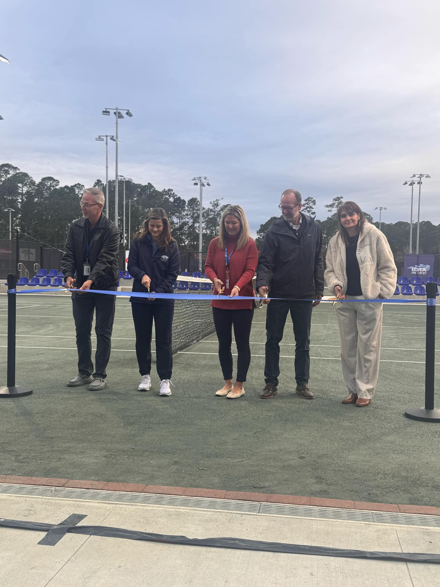 Five people stand on a tennis court, cutting a blue ribbon stretched between two poles Two men and three women are participating in the ceremony, with trees and a cloudy sky in the background