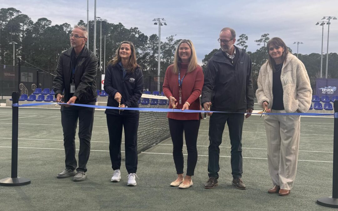 Five people stand on a tennis court holding a large blue ribbon, as if at a ribbon cutting ceremony They are dressed in casual to business casual attire with a net and multiple lights visible in the background