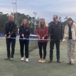 Five people stand on a tennis court holding a large blue ribbon, as if at a ribbon cutting ceremony They are dressed in casual to business casual attire with a net and multiple lights visible in the background