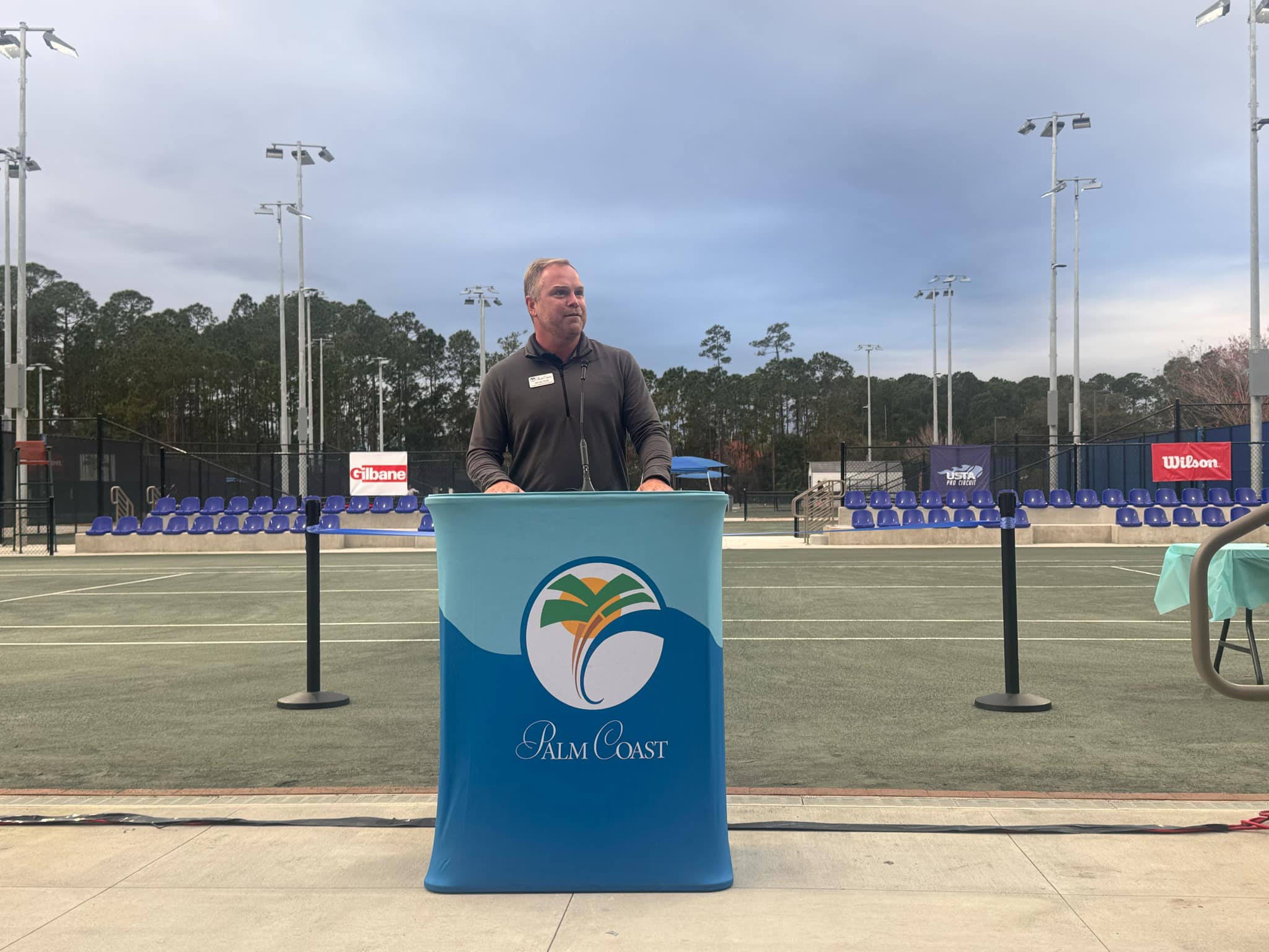 A person stands at a podium with the "Palm Coast" logo, speaking at an outdoor event The setting is a sports court with empty blue seats and banners in the background under a cloudy sky