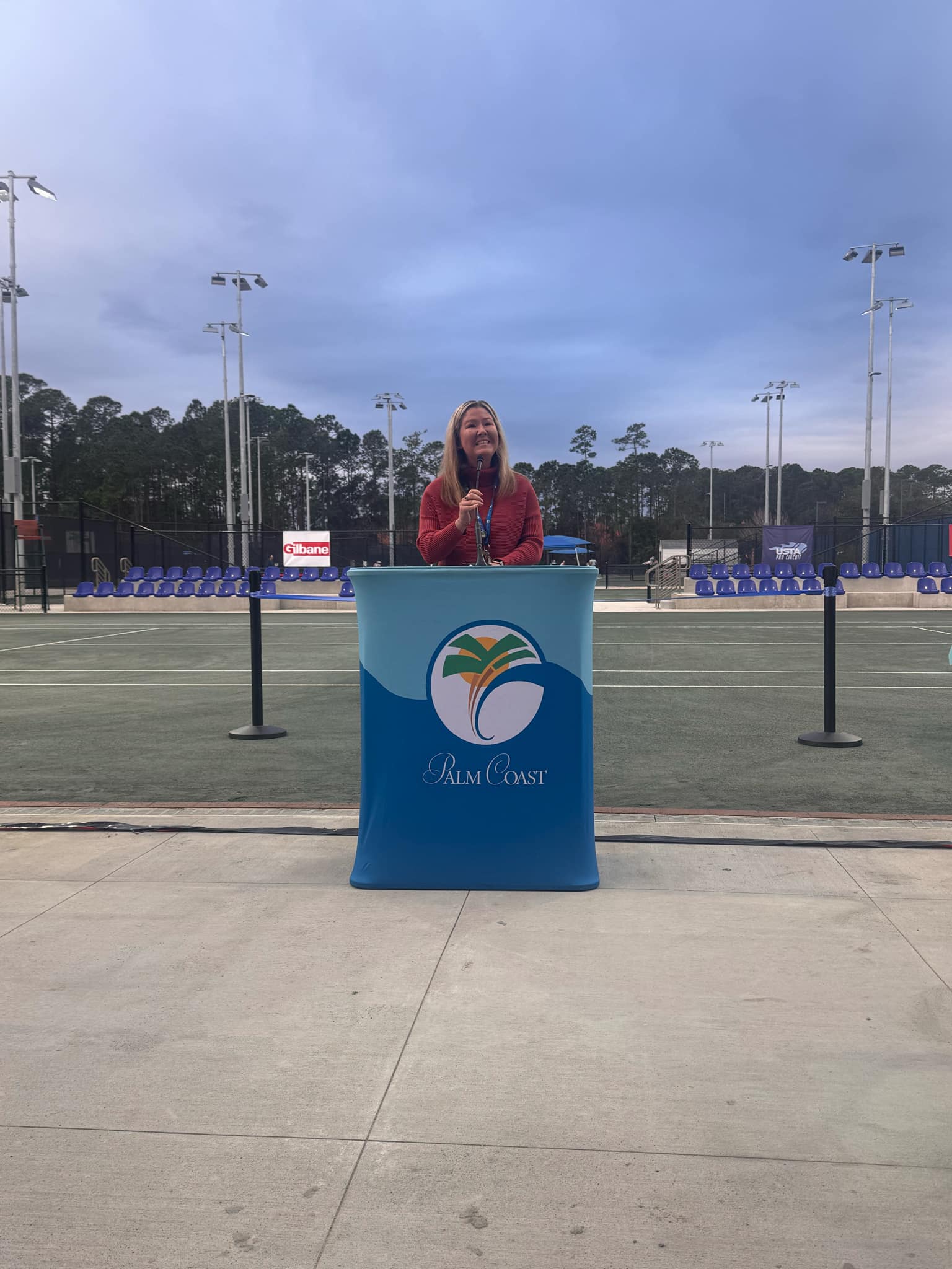 A person stands at a blue podium with a microphone, speaking outdoors on a tennis court The podium displays the Palm Coast logo The background features trees, light poles, and empty seating