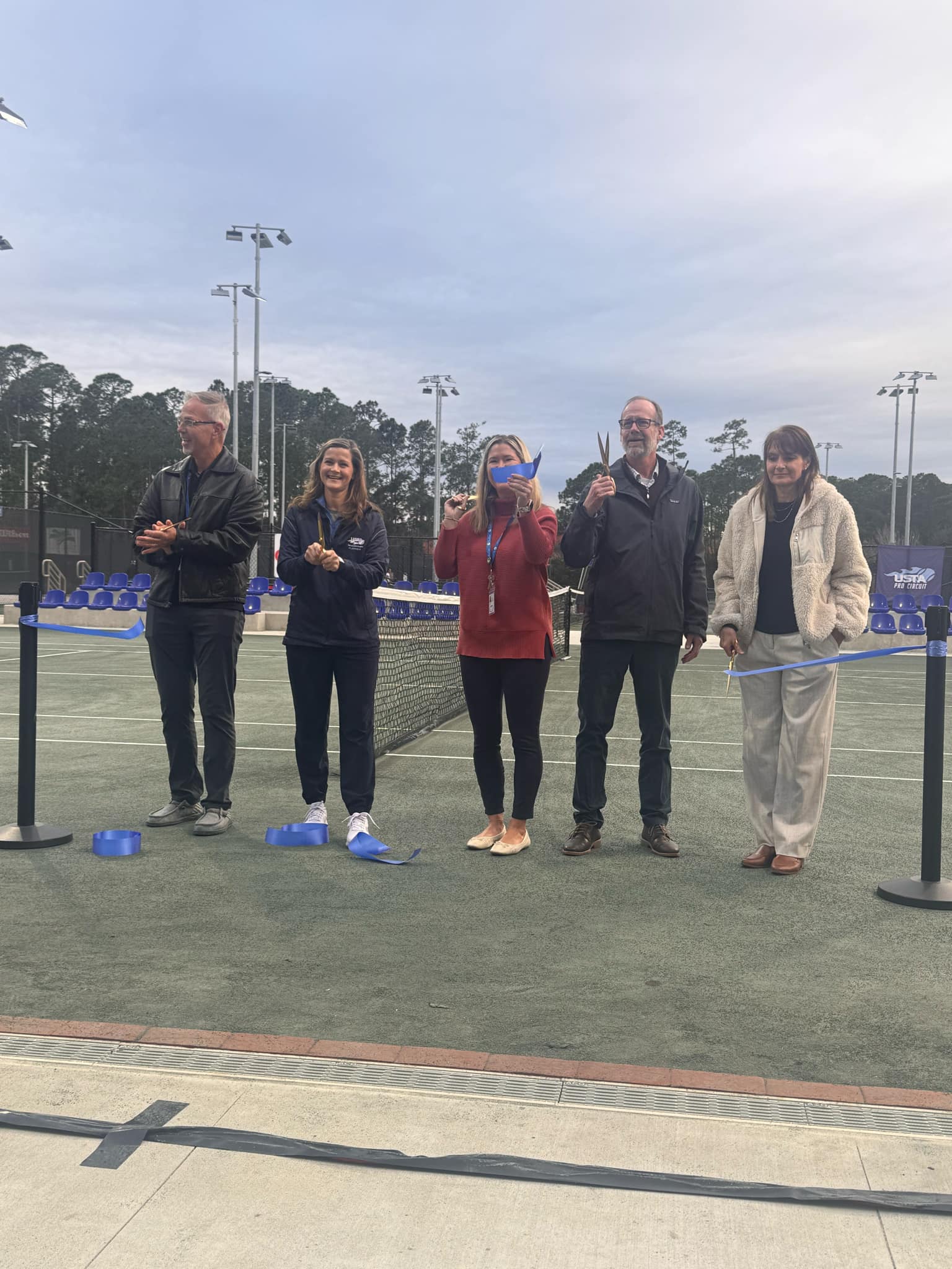 Five people stand on a tennis court, holding large scissors, as if preparing to cut a blue ribbon They are positioned in front of a net with an overcast sky and trees in the background