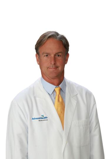 A man wearing a white lab coat with the AdventHealth logo stands against a white background He has short hair and is dressed in a blue shirt and a yellow tie