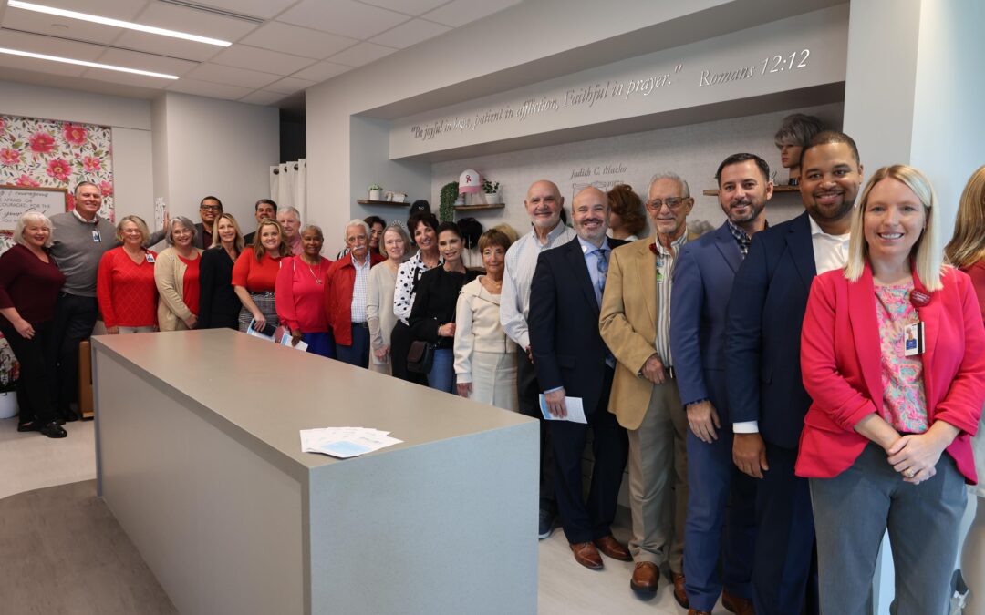 A diverse group of people stand in a well lit room, smiling at the camera They are dressed in business and casual attire A wall in the background displays a decorative design and a quote