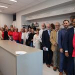 A diverse group of people stand in a well lit room, smiling at the camera They are dressed in business and casual attire A wall in the background displays a decorative design and a quote