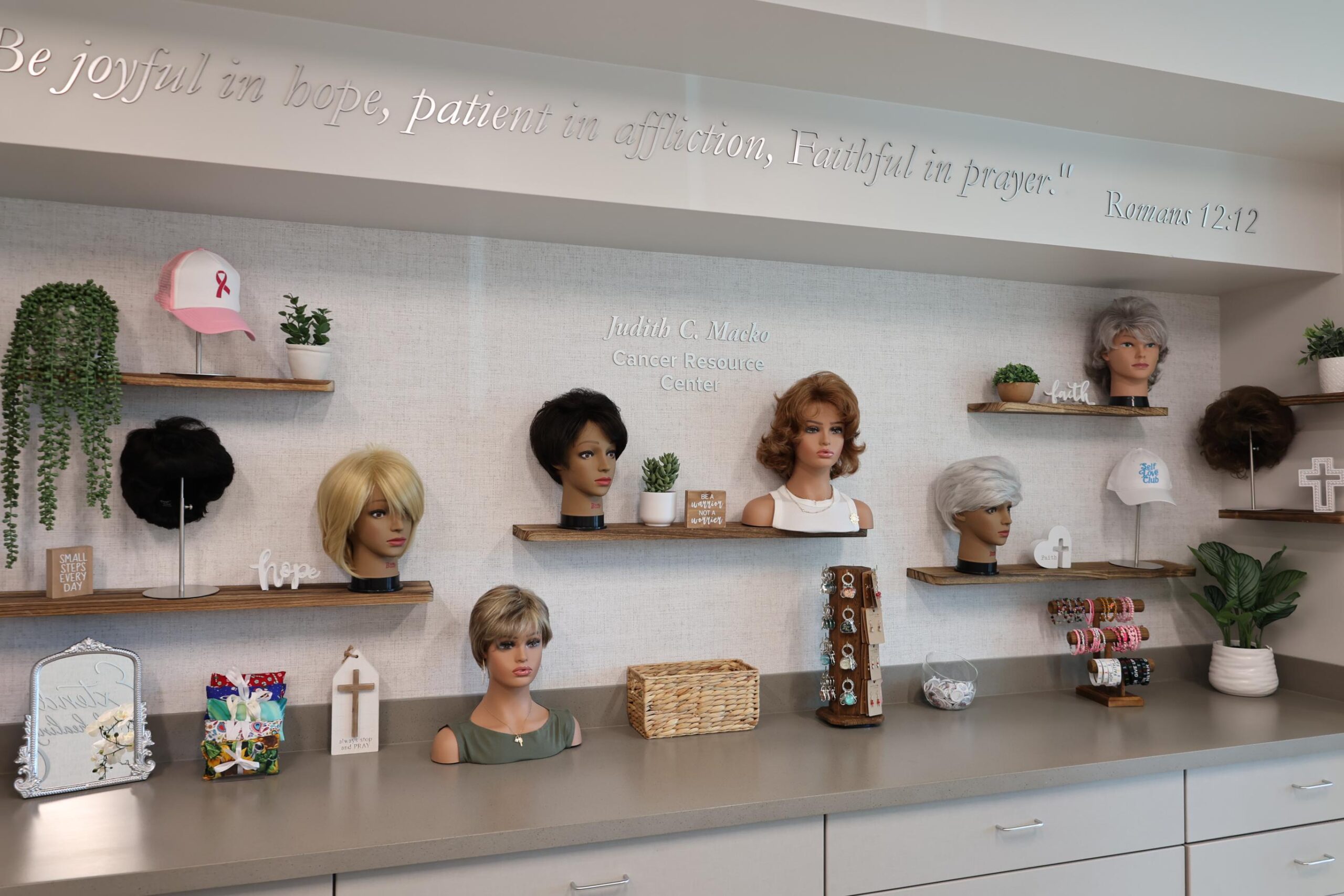 A display of mannequin heads with various wigs on wooden shelves A few decorative items, such as a plant, cross, and encouragement signs, are also present A Bible verse is visible on the wall above: "Be joyful in hope, patient in affliction, faithful in prayer