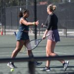 Two women on a tennis court preparing to shake hands over the net One wears a dark tank top and shorts, the other a long sleeve shirt and skirt A tennis ball is in the foreground, and a chain link fence surrounds the court