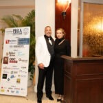 A man in a white tuxedo jacket and a woman in a black dress stand smiling beside a podium A sign displaying various sponsor logos is on their left The setting appears to be a formal event or gathering