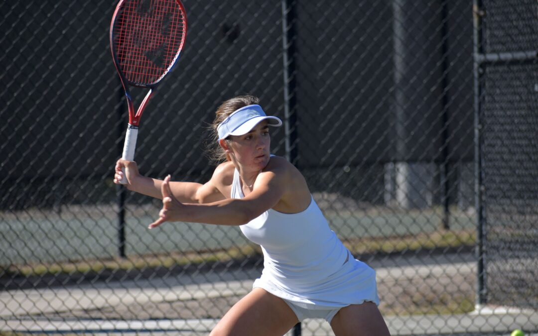 A female tennis player in a white outfit and visor prepares to hit a backhand shot on an outdoor court She holds a red and black tennis racket, with a focused expression The court is surrounded by a chain link fence