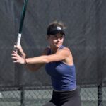 A tennis player in a blue tank top and black visor prepares to hit a backhand shot on an outdoor court The background features a dark chain link fence