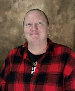 A person wearing a red and black plaid shirt is smiling at the camera They have short hair and hoop earrings The background is a textured brown