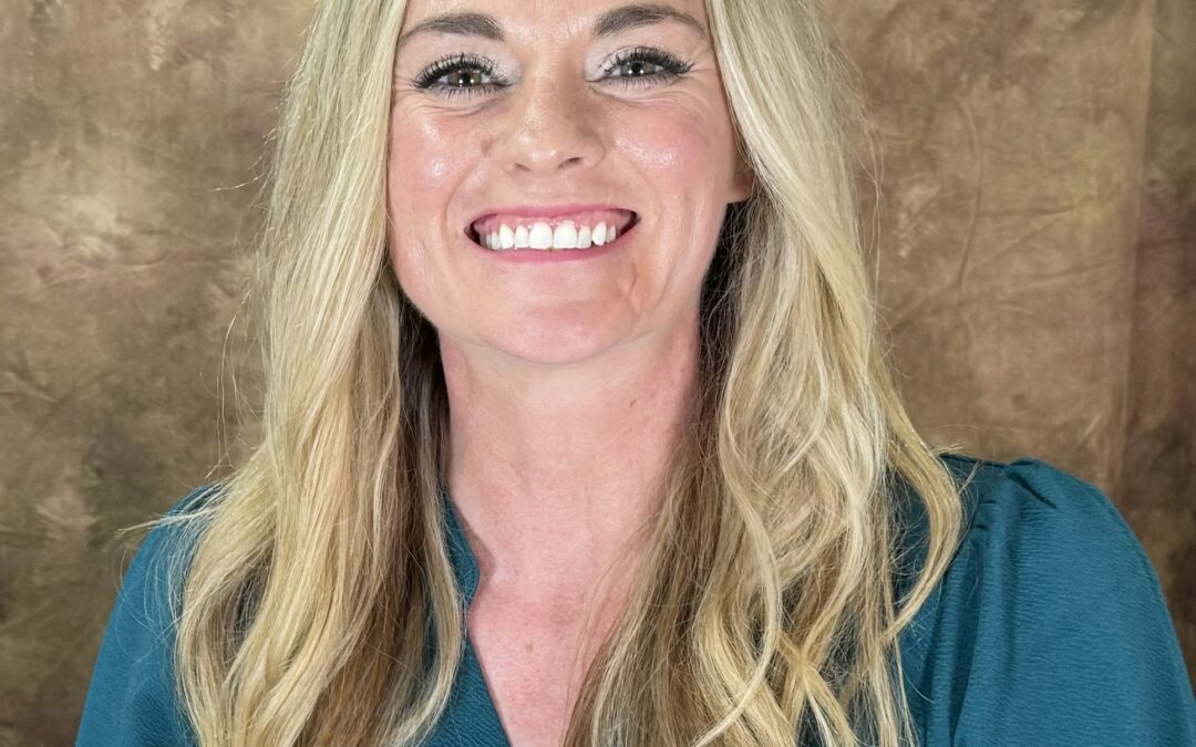 A person with long blonde hair is smiling at the camera, wearing a teal top The background is a textured brown color