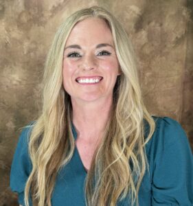 A person with long blonde hair is smiling at the camera, wearing a teal top The background is a textured brown color