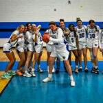 A girls' basketball team poses on a court One player in the center playfully spins a basketball on her finger while teammates stand and cheer behind her A coach stands to the left, smiling, with the team jerseys displaying the number "Vikings