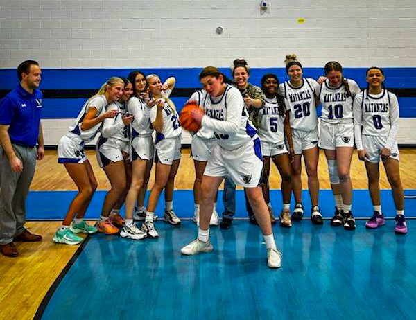 A girls' basketball team poses on a court One player in the center playfully spins a basketball on her finger while teammates stand and cheer behind her A coach stands to the left, smiling, with the team jerseys displaying the number "Vikings