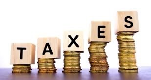 Wooden blocks spelling "TAXES" are placed on stacks of coins of varying heights, suggesting financial growth or burden The background is plain white