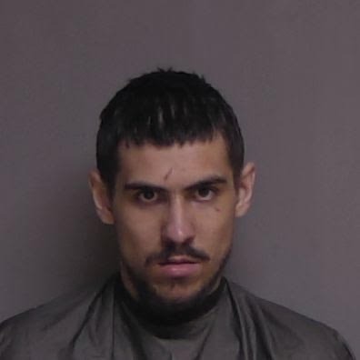 A man with short dark hair, a mustache, and a serious expression is wearing a gray shirt and standing against a plain gray background