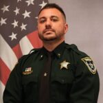 A man in a green sheriff's uniform stands in front of an American flag He has short hair and a beard and wears a badge and insignia on his uniform