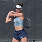 A woman in sportswear and a visor is holding a tennis racket, preparing to swing She stands in front of a chain link fence with a focused expression