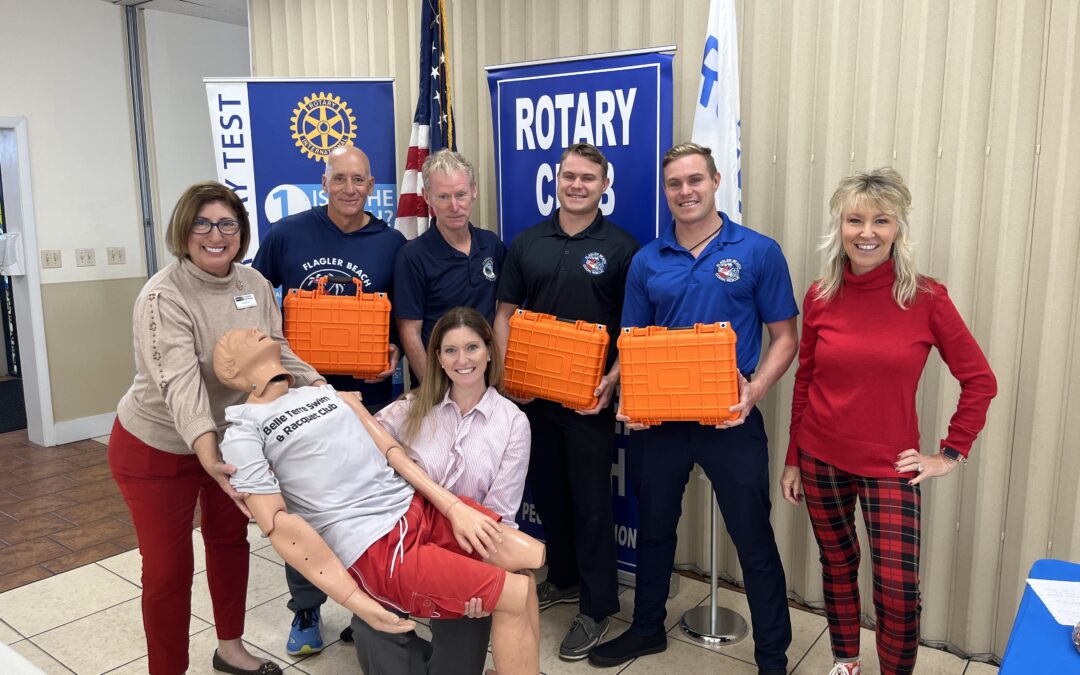 A group of people stand together indoors, holding orange boxes with a CPR training dummy Banners with "Rotary Club" are visible in the background Some are dressed casually, while others wear uniforms