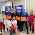 A group of people stand together indoors, holding orange boxes with a CPR training dummy Banners with "Rotary Club" are visible in the background Some are dressed casually, while others wear uniforms