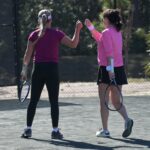 Two women on a tennis court share a fist bump Both hold rackets; one wears a purple top and black leggings, the other wears a pink top and black skirt The court is outdoors with a chain link fence and trees in the background