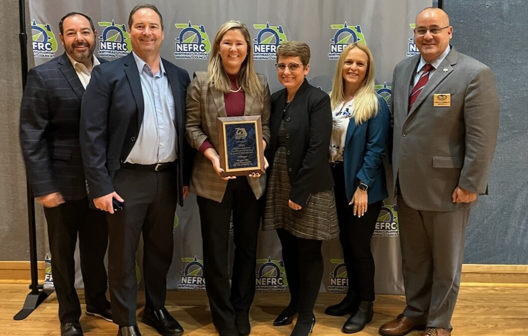 A group of six people smiling and standing in front of a NEFRC banner One person in the middle holds a plaque They are dressed in formal attire, and the event appears to be an award ceremony