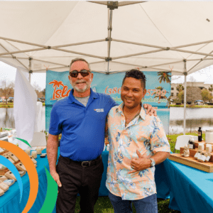 Two men stand smiling under a white canopy at an outdoor event The man on the left wears sunglasses and a blue polo shirt, while the man on the right wears a floral shirt A colorful table with products and a scenic banner are behind them