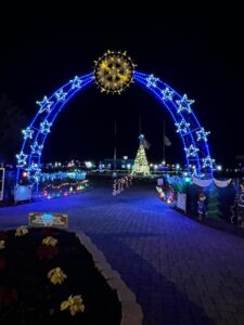 A nighttime scene features a large arch adorned with blue and white lights, topped by a star shaped decoration In the background, a brightly lit Christmas tree stands, surrounded by festive lights and decorations A paver path leads under the arch