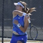 A tennis player wearing a blue outfit and white visor is hitting a forehand shot She has a focused expression and is on an outdoor court, with a chain link fence in the background Her hair is in a ponytail