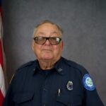 An older person wearing a dark blue uniform poses for a portrait, standing in front of an American flag The uniform has various patches and insignia on the sleeves and chest The background is a neutral gray