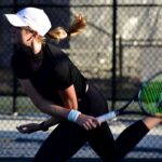 A female tennis player in a black outfit and white cap is in motion on the court, swinging a racket The background features a chain link fence Her ponytail is flying, and she wears a white wristband