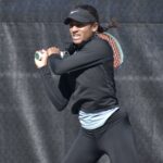 A person in athletic wear prepares to hit a two handed backhand in tennis They're wearing a black visor and long sleeve top, with a tennis racket held behind them The background is a dark chain link fence