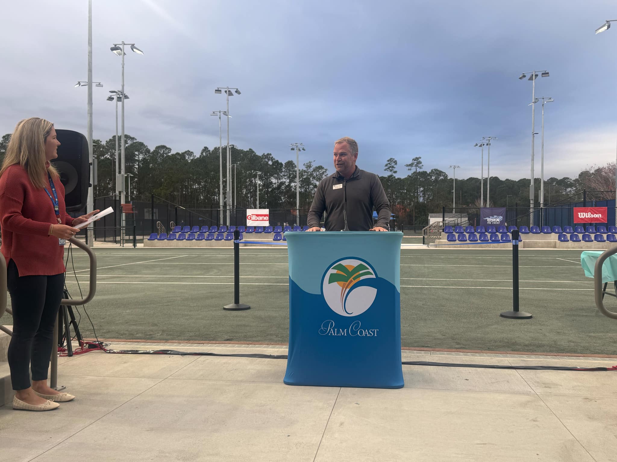 A man speaks at a podium with the Palm Coast logo at an outdoor tennis court A woman stands nearby holding papers There are tennis balls and nets in the background under a cloudy sky