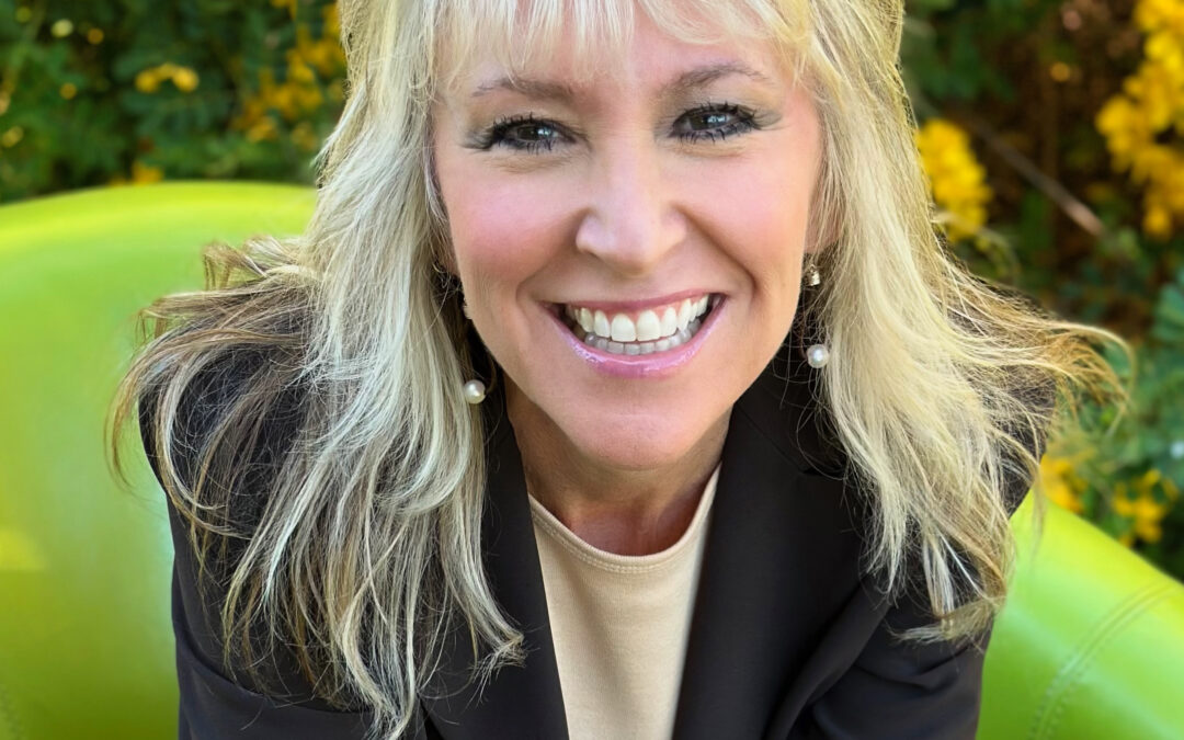 A smiling woman with long blonde hair is sitting in a green chair She is wearing a black blazer over a beige top Behind her, there is a backdrop of bright yellow flowers