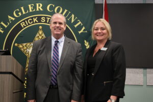 Two people stand smiling in front of a green banner featuring the words "Flagler County" and a star emblem The man is wearing a suit and striped tie, while the woman is dressed in a black blazer