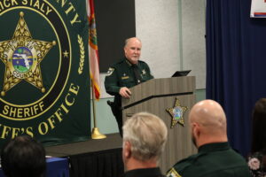 A sheriff in uniform stands at a podium giving a speech A large sheriff's office emblem and a Florida flag are displayed in the background Several people, also in uniform, are seated and facing the speaker